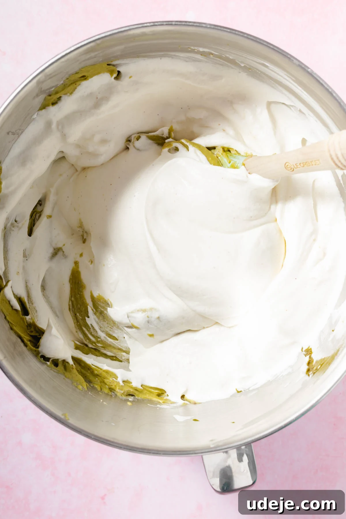 Lush Eggless Pistachio Tiramisu 11 A metal mixing bowl filled with fluffy, whipped cream mixture for pistachio tiramisu is being stirred with a wooden spatula, set against a light pink background.