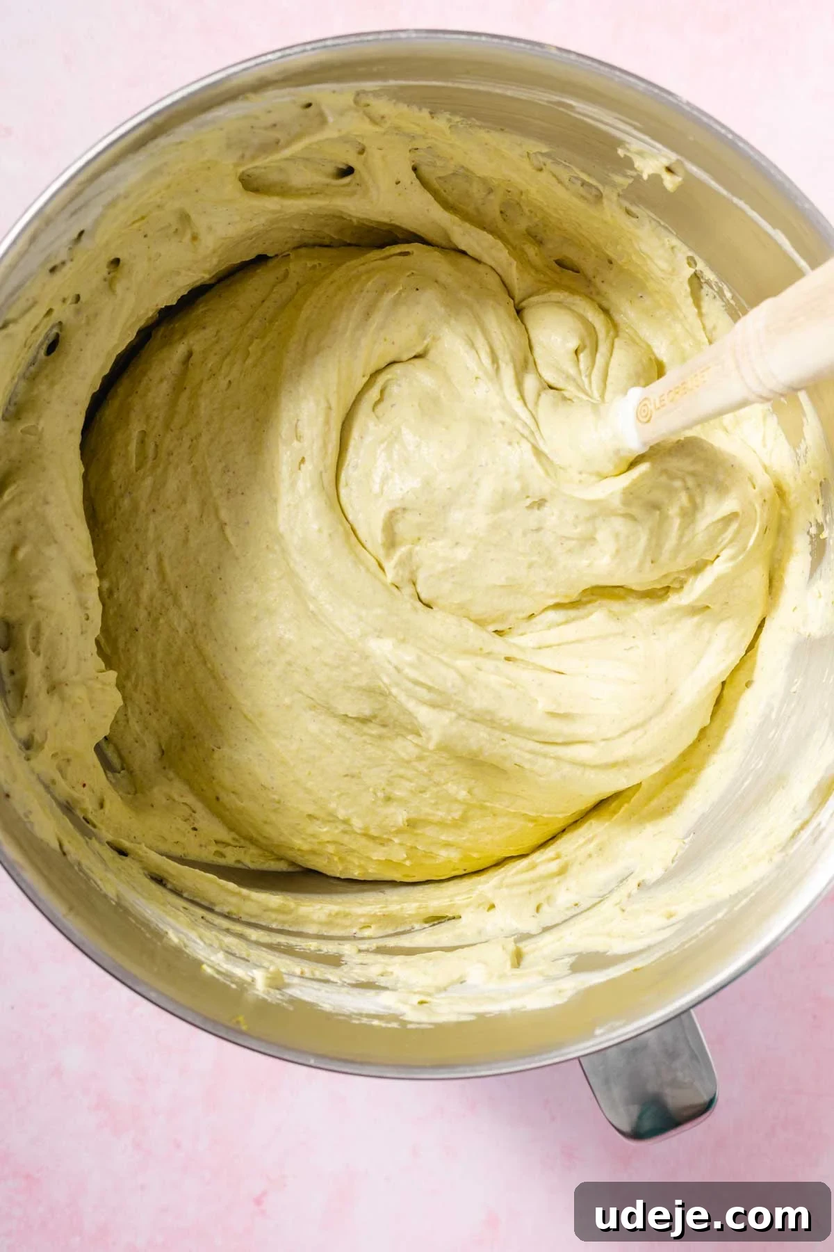 Lush Eggless Pistachio Tiramisu 12 A mixing bowl filled with thick, creamy pistachio tiramisu cake batter being stirred with a wooden spatula, set on a light pink surface.