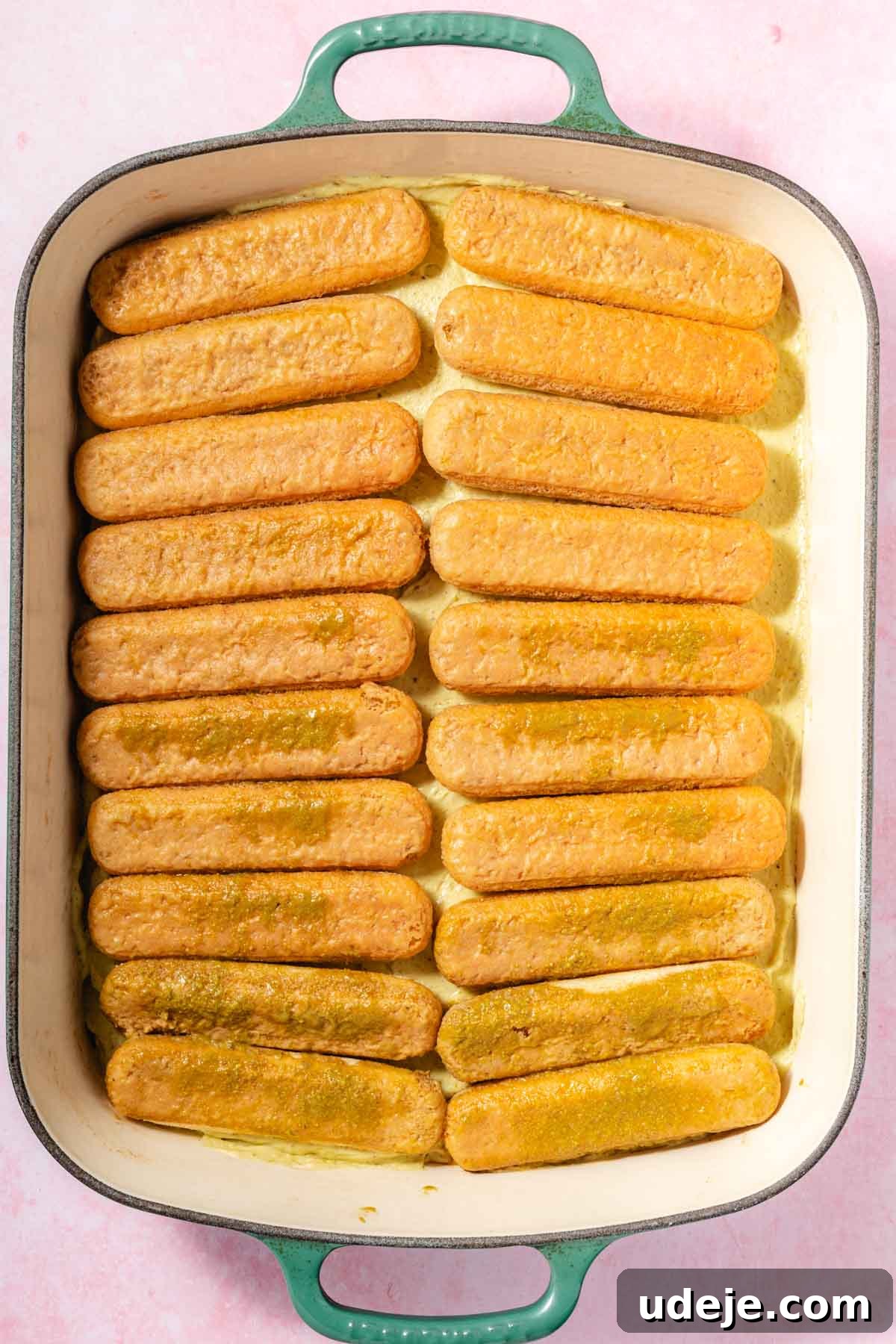 Lush Eggless Pistachio Tiramisu 13 A baking dish filled with two layers of ladyfinger cookies soaked in a light yellow liquid, arranged neatly in rows—this pistachio tiramisu rests in a dish with green handles on a pale pink surface.
