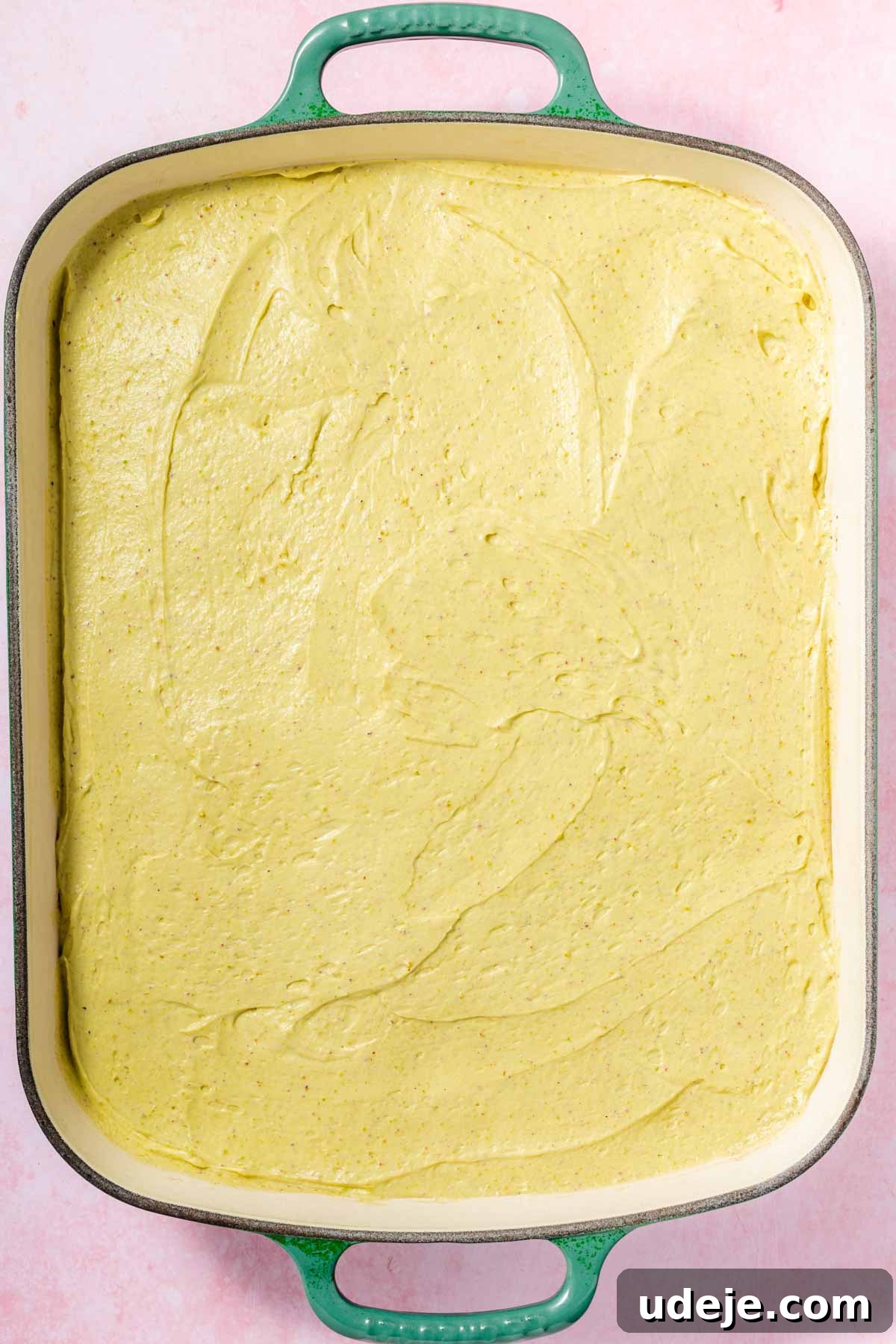 Lush Eggless Pistachio Tiramisu 14 A large rectangular baking dish with green handles contains a smooth layer of yellow cornbread batter, ready to be baked—reminiscent of the layers found in pistachio tiramisu. The dish sits on a light pink surface.