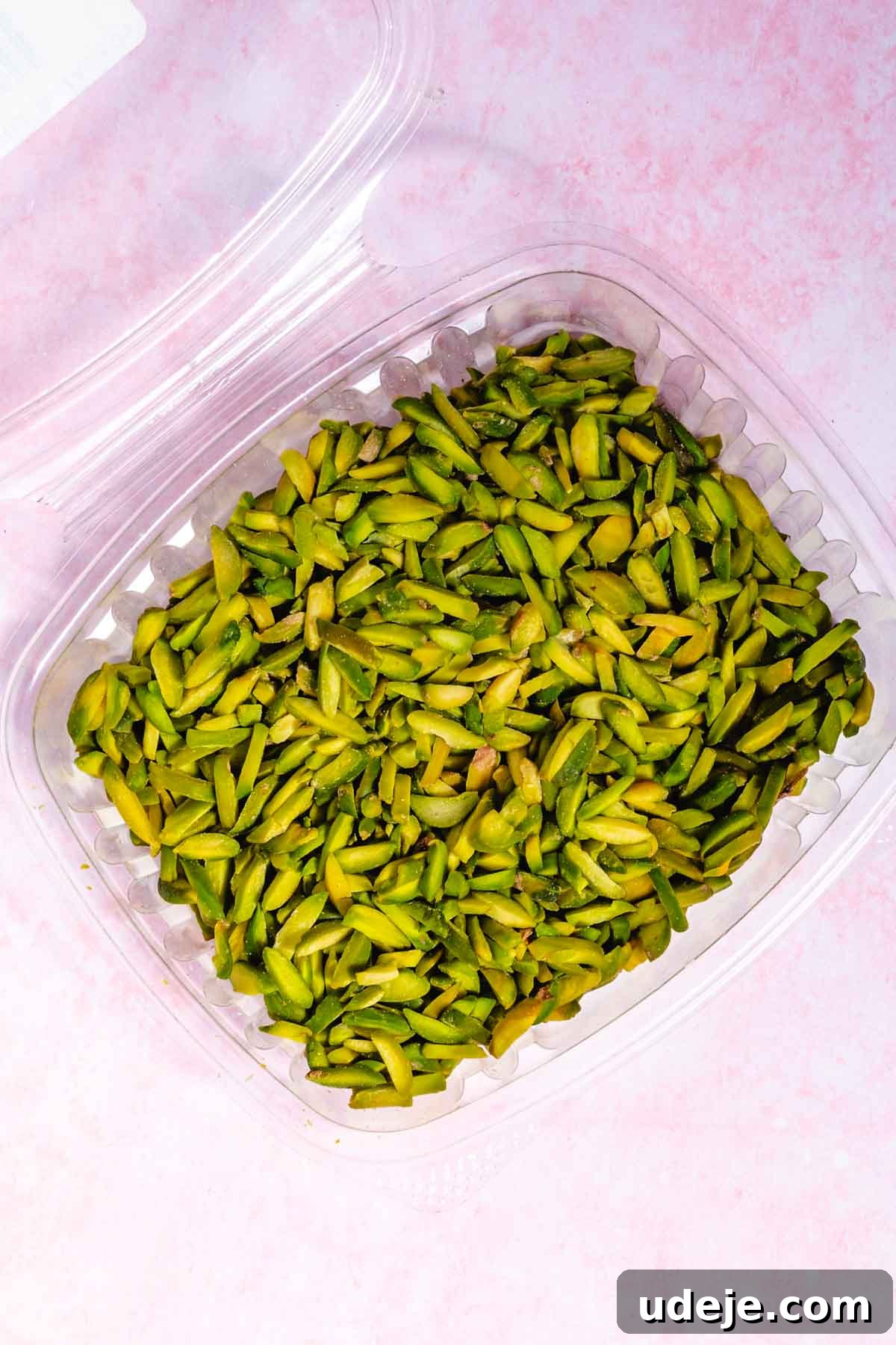 Lush Eggless Pistachio Tiramisu 15 A clear plastic container filled with sliced green pistachios, perfect for topping your homemade pistachio tiramisu, sits on a light pink surface.