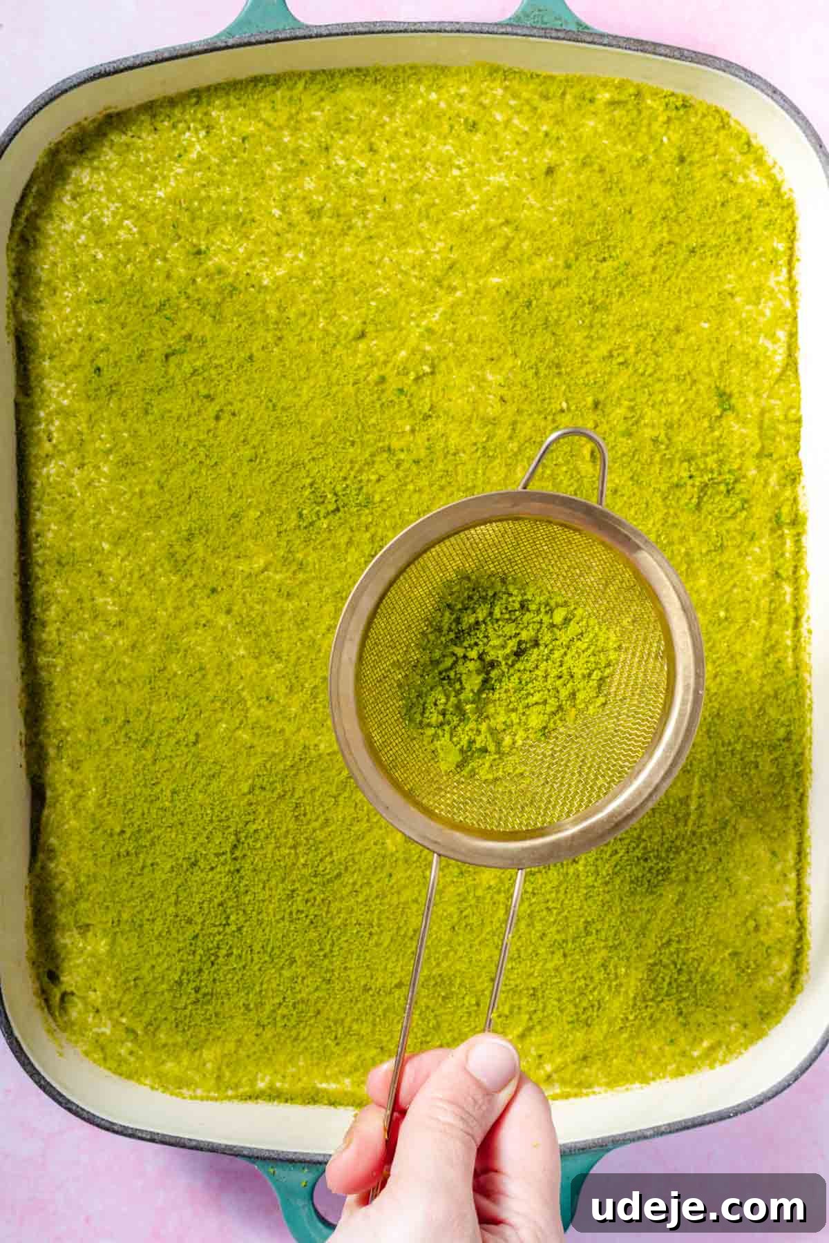 Lush Eggless Pistachio Tiramisu 17 A hand holds a fine mesh sieve with green matcha powder, gently dusting the surface of a large baking dish filled with smooth pistachio tiramisu, creating an even, elegant finish.