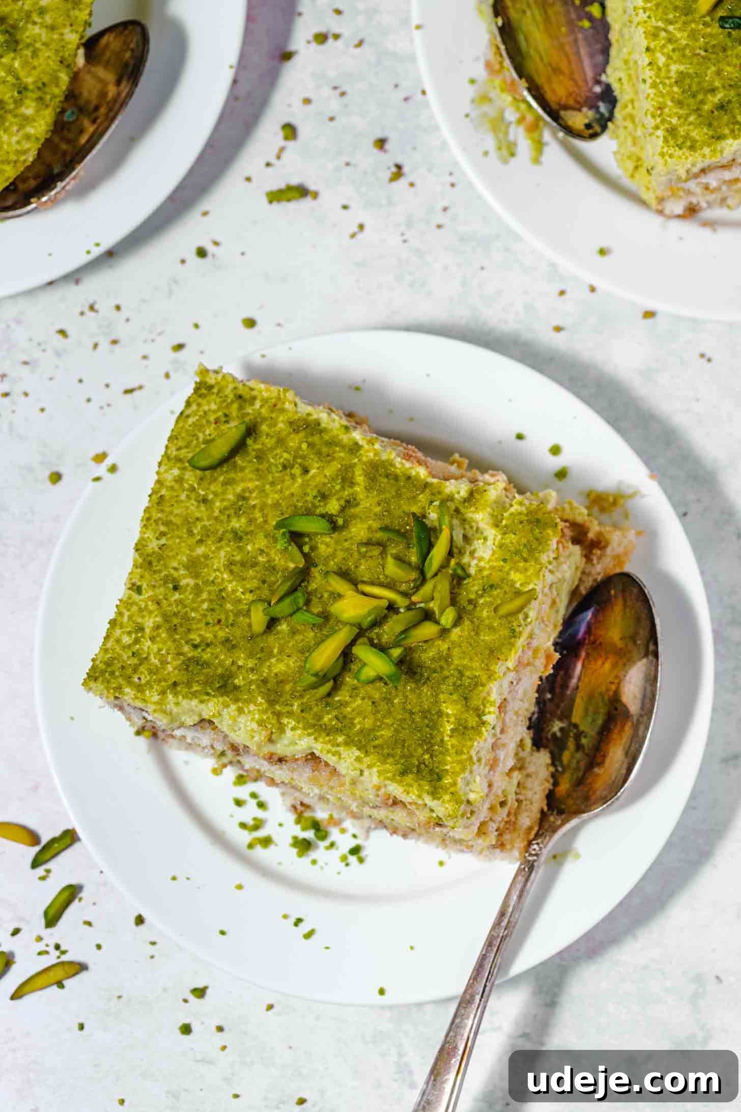 Lush Eggless Pistachio Tiramisu 18 A square slice of pistachio tiramisu with green topping and chopped pistachios sits on a white plate next to a spoon. The plate rests on a white surface, accented by scattered crumbs and slivers of pistachio.