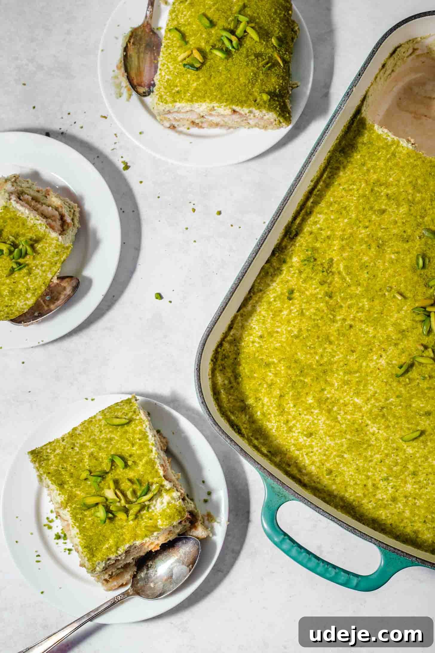 Lush Eggless Pistachio Tiramisu 3 A rectangular dish filled with pistachio tiramisu. Two square slices are served on white plates, each garnished with chopped pistachios. A spoon rests on one plate. The scene is bright and top-lit.