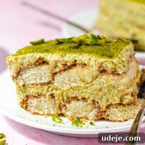 A slice of pistachio tiramisu with green pistachio topping is served on a white plate with a fork, highlighting the creamy layers. In the background, another portion of this delightful pistachio tiramisu sits on a pink surface.
