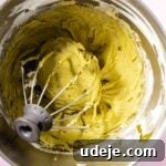 Lush Eggless Pistachio Tiramisu 21 Overhead view of a metal mixing bowl with a wire whisk attachment and thick, greenish-yellow pistachio tiramisu batter inside, set against a pale pink background.