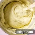 Lush Eggless Pistachio Tiramisu 22 A metal mixing bowl filled with thick, creamy cake batter for pistachio tiramisu. A wooden spatula is partially immersed in the pale yellow batter. The bowl rests on a light pink surface.