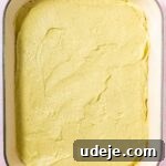 Lush Eggless Pistachio Tiramisu 24 A large rectangular baking dish with green handles containing an even layer of yellow cake batter, ready to be baked for a delightful pistachio tiramisu. The background is a light pink surface.