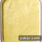 Lush Eggless Pistachio Tiramisu 25 A rectangular green baking dish filled with an even layer of yellow cake batter, ready to be baked. The creamy batter is smooth with visible spatula marks, reminiscent of layers in a pistachio tiramisu.