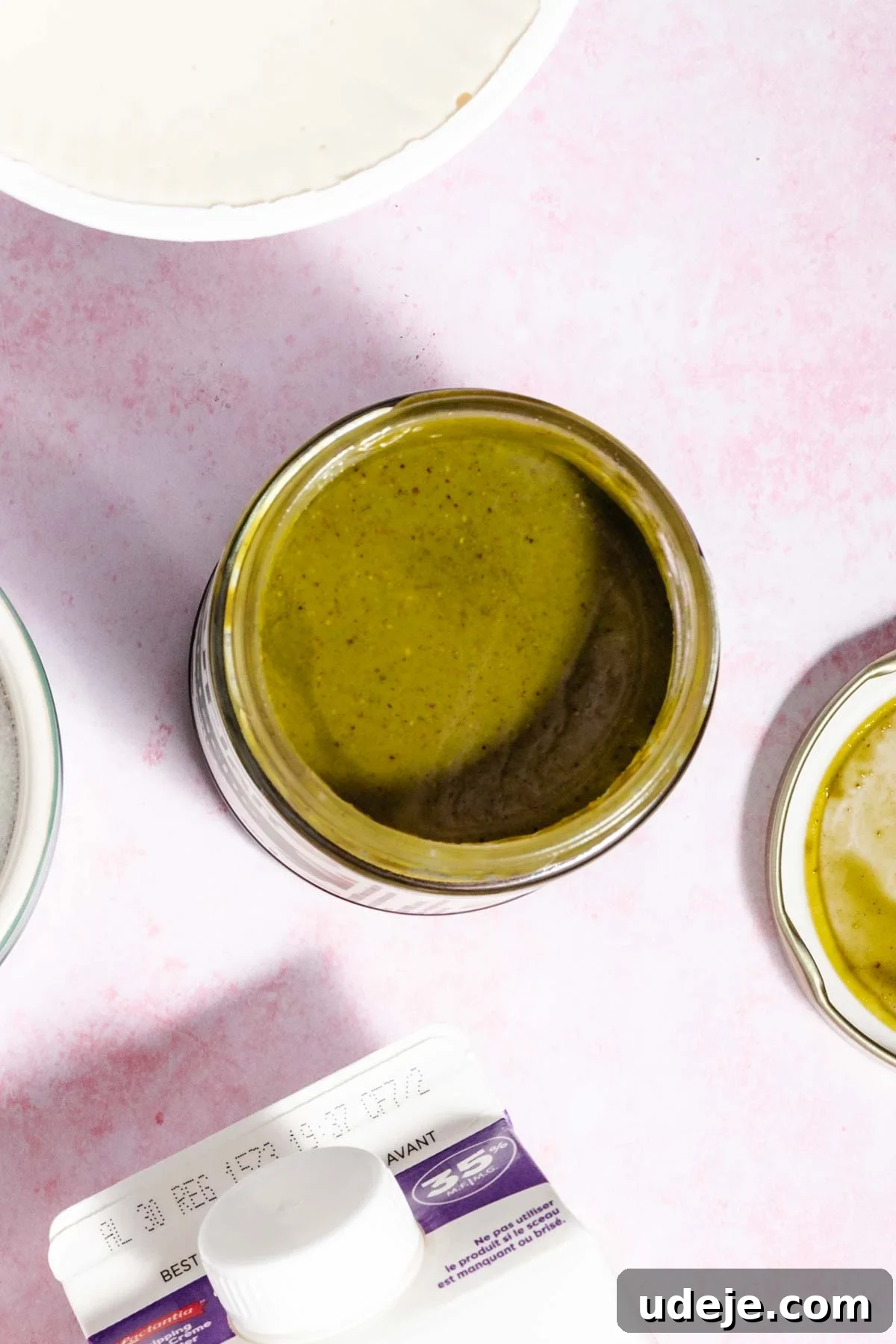 Lush Eggless Pistachio Tiramisu 4 A jar filled with green sauce sits on a light pink surface, surrounded by a white dish, a small bowl with more sauce, and the corner of a carton—perfect for drizzling over pistachio tiramisu.
