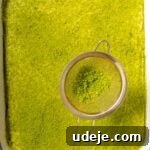 Lush Eggless Pistachio Tiramisu 27 A hand holds a small sieve, sprinkling matcha powder over a large rectangular dish filled with creamy green pistachio tiramisu, set against a pink background.