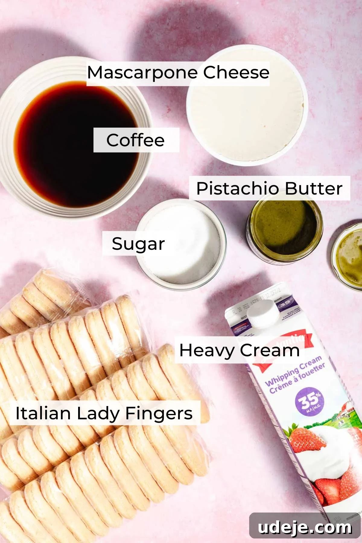 Lush Eggless Pistachio Tiramisu 5 Top-down view of ingredients for pistachio tiramisu: mascarpone cheese, coffee, pistachio butter, sugar, heavy cream, and Italian lady fingers, all labeled and arranged on a pink surface.