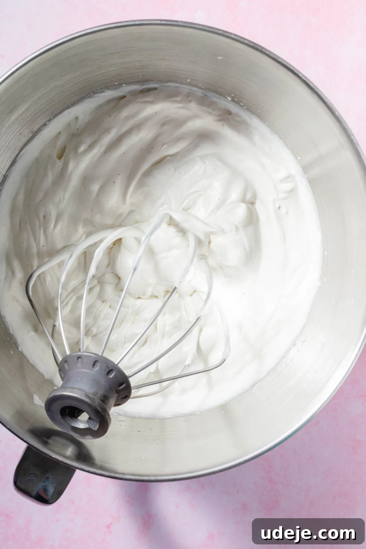 Lush Eggless Pistachio Tiramisu 7 A metal whisk attachment rests in a mixing bowl filled with thick, freshly whipped cream, ready to be layered into a decadent pistachio tiramisu, set on a light pink surface.
