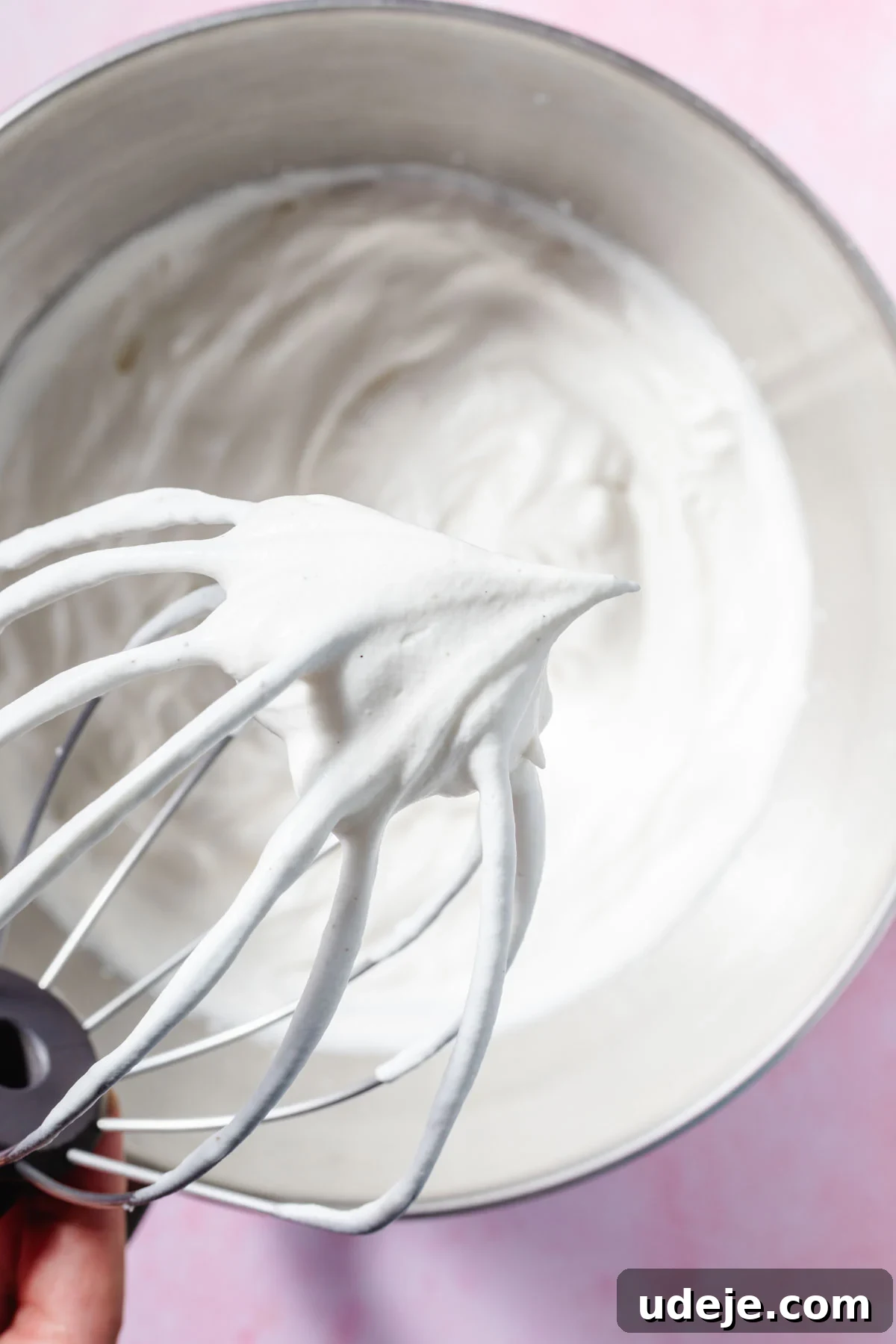 Lush Eggless Pistachio Tiramisu 8 A close-up of a metal whisk with stiff, glossy whipped cream peaks, held above a mixing bowl filled with more whipped cream for pistachio tiramisu, on a light pink surface.
