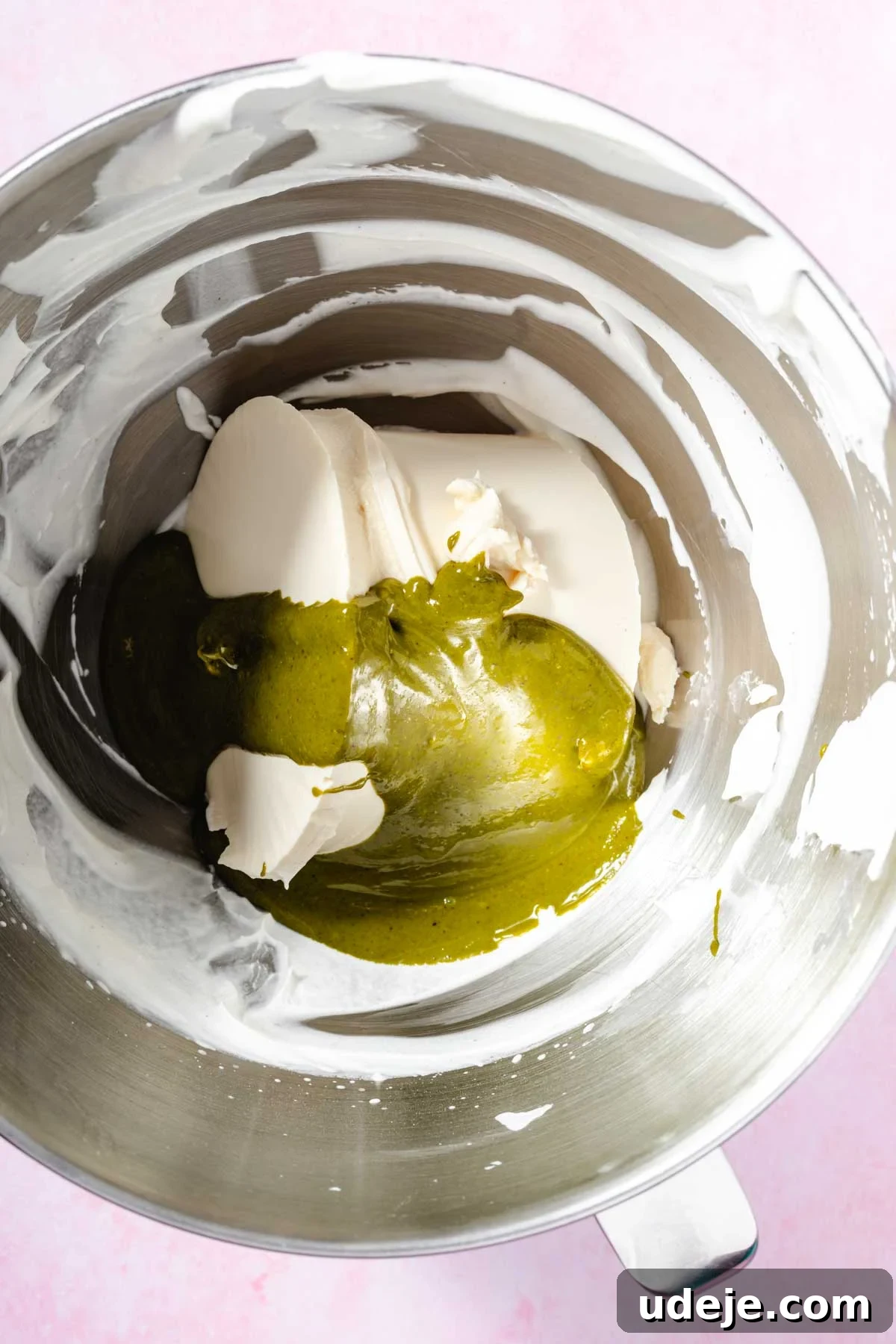 Lush Eggless Pistachio Tiramisu 9 A metal mixing bowl containing partially mixed white cream cheese and a green matcha mixture for pistachio tiramisu, set against a light pink background.
