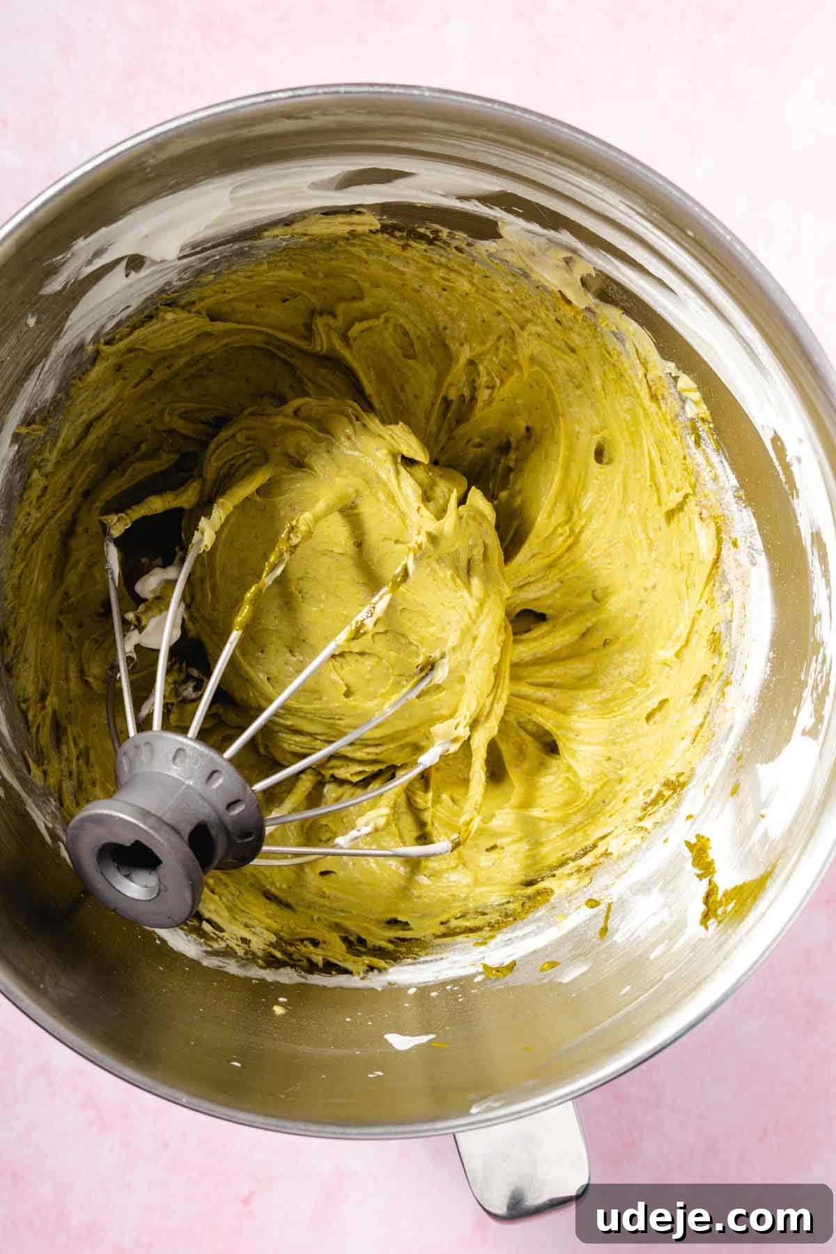 Lush Eggless Pistachio Tiramisu 10 Overhead view of a metal mixing bowl containing thick matcha green tea batter, partially mixed with a whisk attachment, evokes the creamy texture found in pistachio tiramisu, against a light pink background.