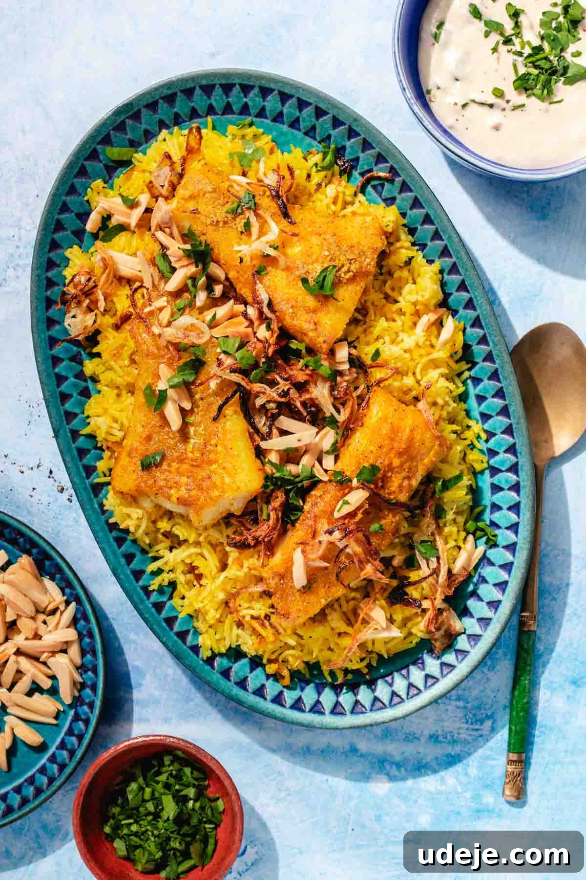 Fragrant Middle Eastern Fish Pilaf 2 Blue patterned platter with yellow rice, fried cod fillets, garnished with toasted slivered almonds, onions and parsley, with a metal spoon beside it, a small blue patterned bowl with toasted almonds, a small orange bowl with parsley and a blue bowl with tahini sauce on a blue background