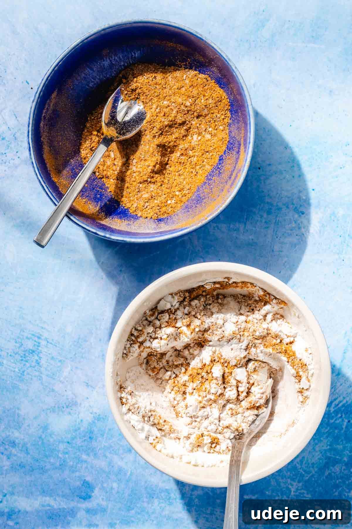 Fragrant Middle Eastern Fish Pilaf 11 Mixed spice in a blue bowl with a metal spoon beside a white bowl with some spices and cornstarch and a metal spoon on a blue background