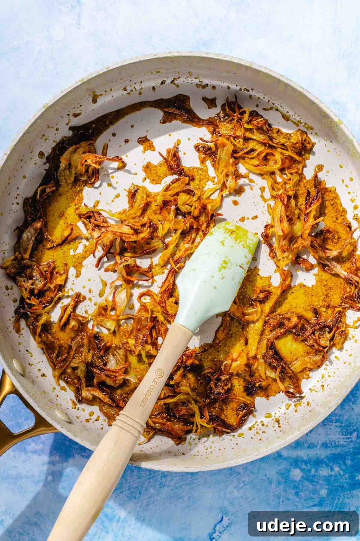 Fragrant Middle Eastern Fish Pilaf 17 crispy onions being fried with spices in a saute pan with a blue spatula on a blue background
