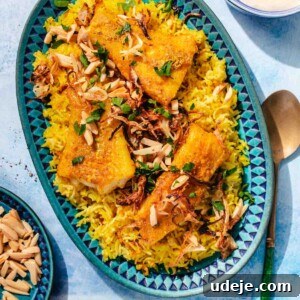 Fragrant Middle Eastern Fish Pilaf 20 Blue patterned platter with yellow rice, fried cod fillets, garnished with toasted slivered almonds, onions and parsley, with a metal spoon beside it, a small blue patterned bowl with toasted almonds, a small orange bowl with parsley and a blue bowl with tahini sauce on a blue background