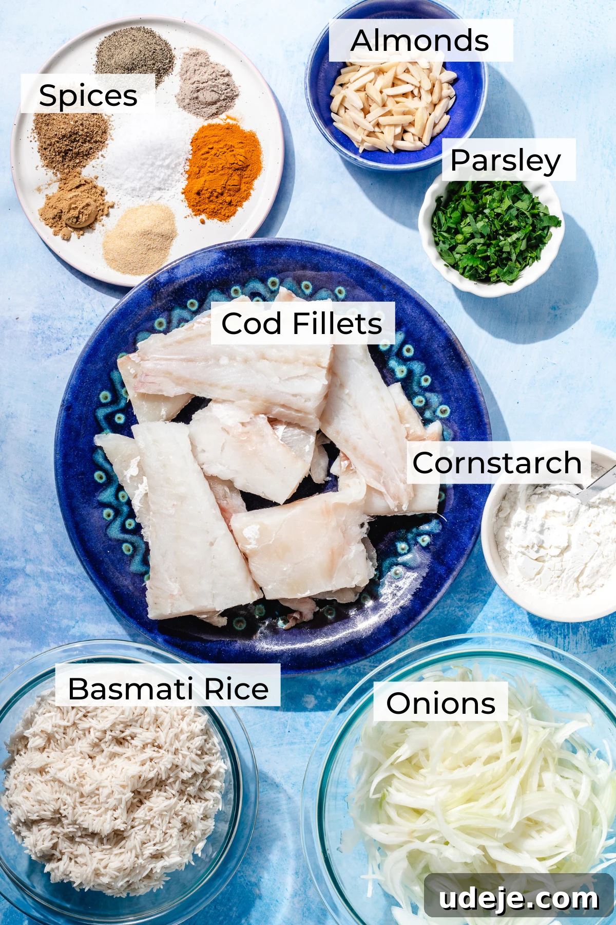 Fragrant Middle Eastern Fish Pilaf 3 Ingredient shot for sayadieh with a white plate with spices, slivered almonds in a blue bowl, chopped parsley in a white bowl, a blue patterned plate with cod fillets, a white bowl with cornstarch, a glass bowl with basmati rice, a glass bowl with sliced onions on a blue background