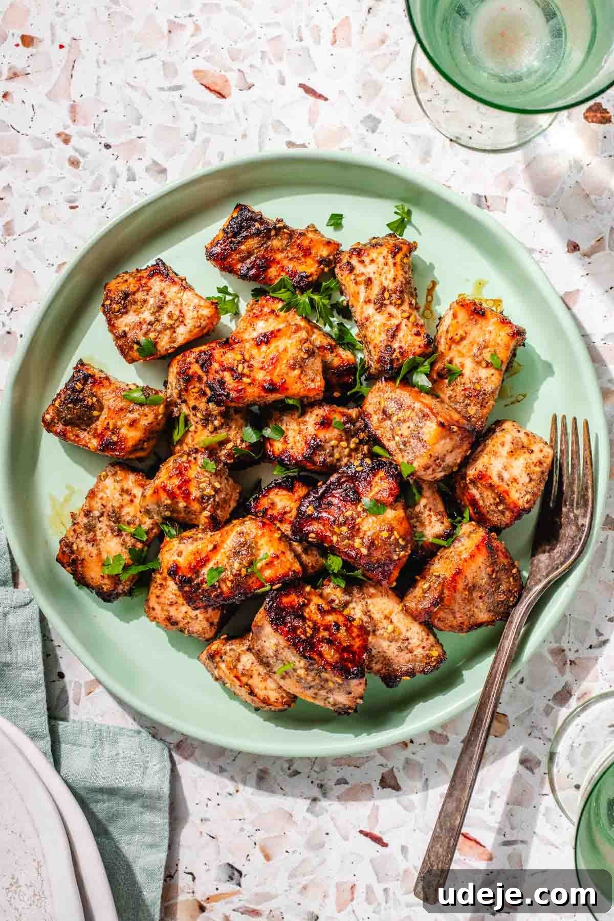 Crispy Air Fried Zaatar Salmon Bites 2 Crispy Air Fryer Salmon Bites garnished with fresh parsley on a light green plate, set against a green tea towel with a glass of water and a metal fork, all on a pink terrazzo background.