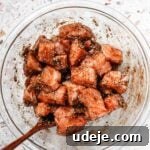 Cubed salmon gently mixed with marinade in a glass bowl using a wooden spoon on a pink terrazzo background.