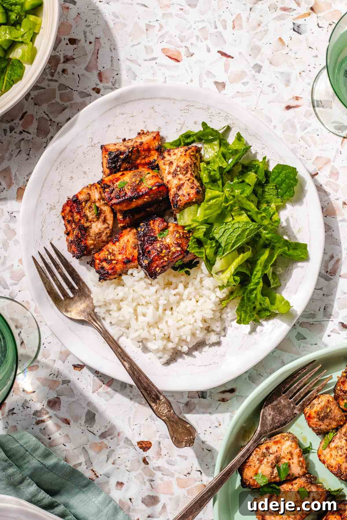 Crispy Air Fried Zaatar Salmon Bites 3 A white plate featuring fluffy rice, perfectly cooked air fryer salmon bites, and fresh lettuce, alongside a green plate with more salmon bites and a metal fork, all on a vibrant pink terrazzo background.
