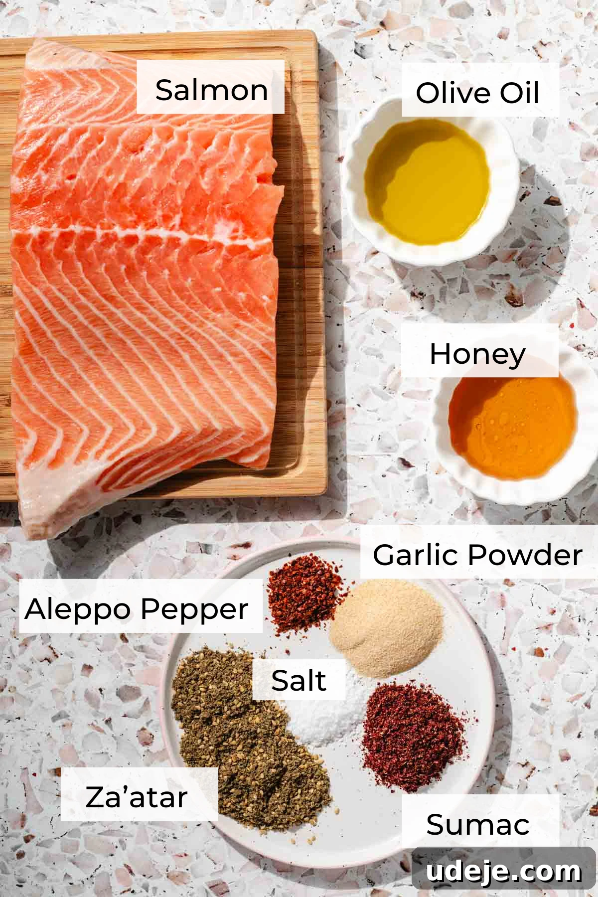 Crispy Air Fried Zaatar Salmon Bites 4 A selection of ingredients for air fryer salmon: fresh salmon on a wooden cutting board, small white bowls containing olive oil and honey, and a white plate holding a blend of spices, all arranged on a pink terrazzo background.