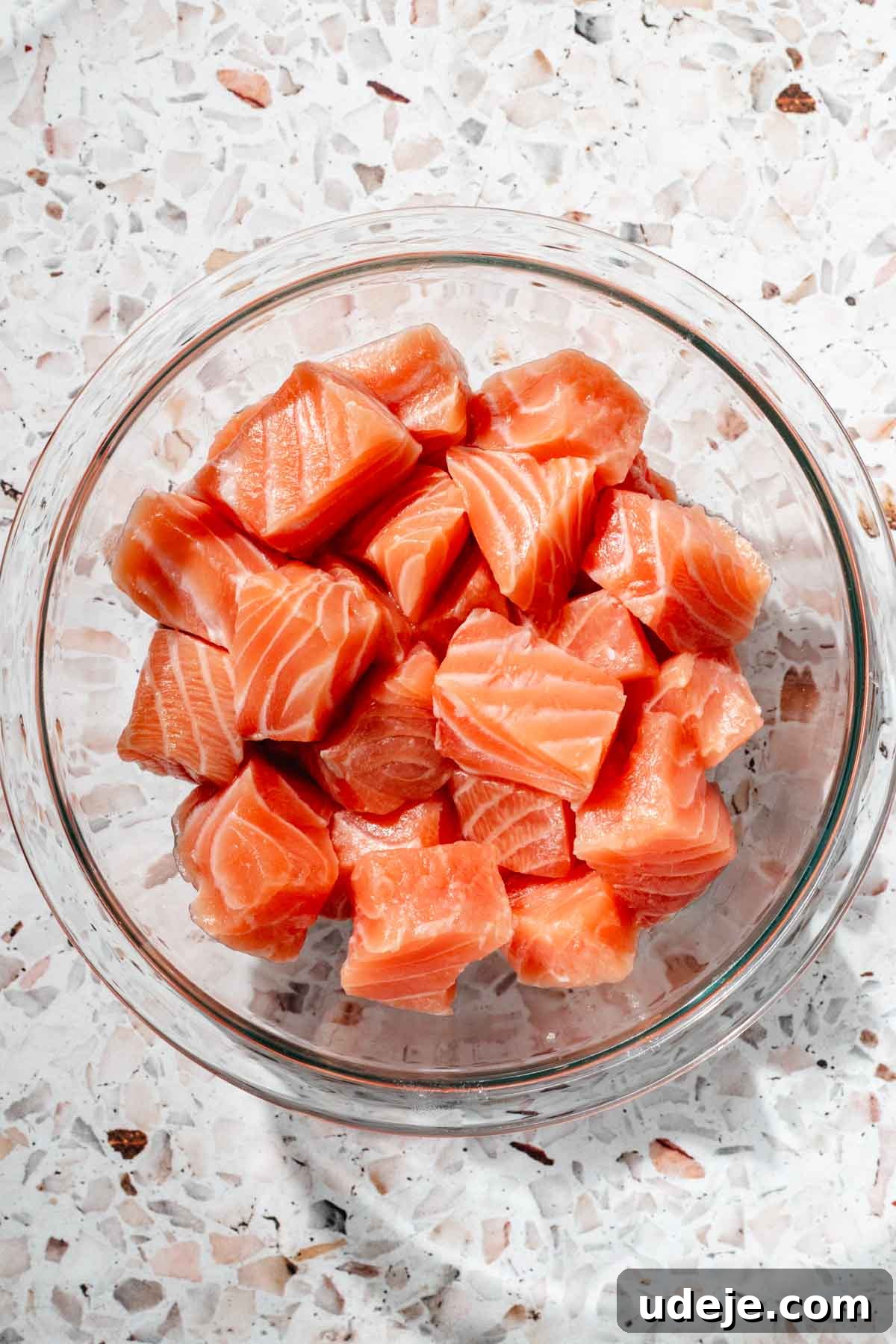 Crispy Air Fried Zaatar Salmon Bites 5 Uniformly cubed salmon in a clear glass bowl, ready for marinating, displayed on a pink terrazzo background.