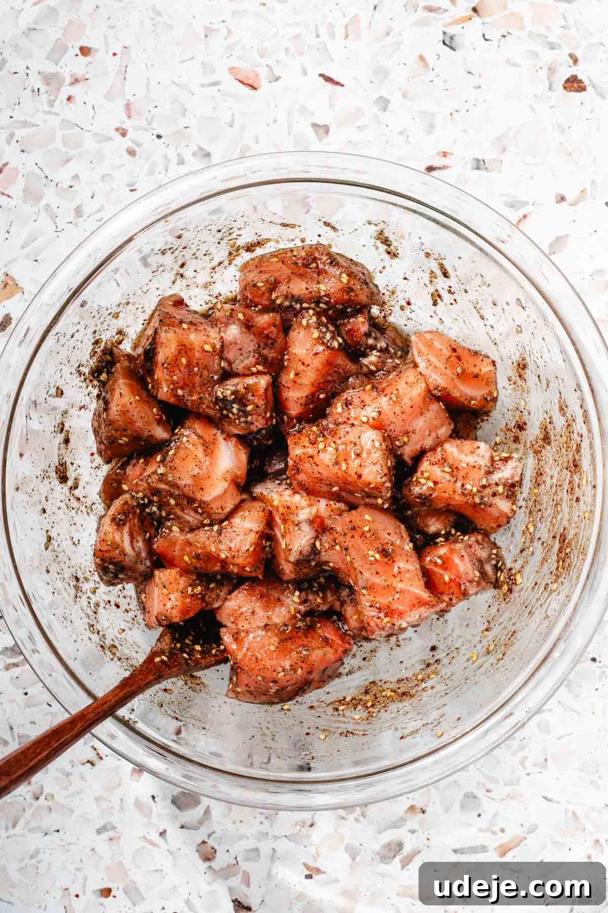 Crispy Air Fried Zaatar Salmon Bites 7 Cubed salmon, thoroughly mixed with a rich marinade in a glass bowl, with a small wooden spoon, all on a pink terrazzo background.