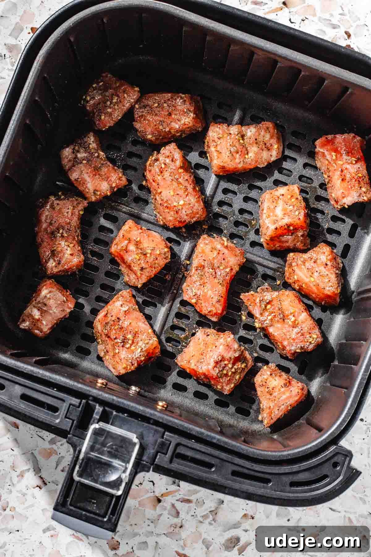 Crispy Air Fried Zaatar Salmon Bites 8 Raw, marinated, cubed salmon neatly arranged in a black air fryer basket, ready for cooking, set on a pink terrazzo background.