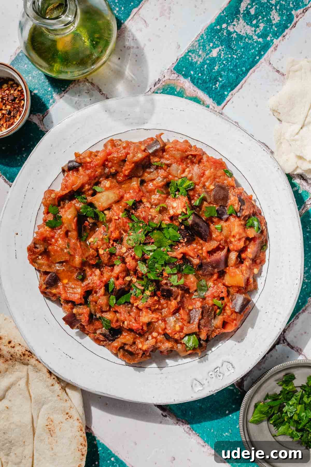 Speedy Moroccan Eggplant Zaalouk 2 Zaalouk on a plate, garnished with parlsey with pita next to it, a small bowl with chilli flakes, a small jug of olive oil, a small plate of chopped parsley on a green and white tiled background