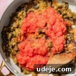 Speedy Moroccan Eggplant Zaalouk 8 Grated tomatoes added to cooked, smashed eggplant in a saute pan with a wooden spoon on a pink background