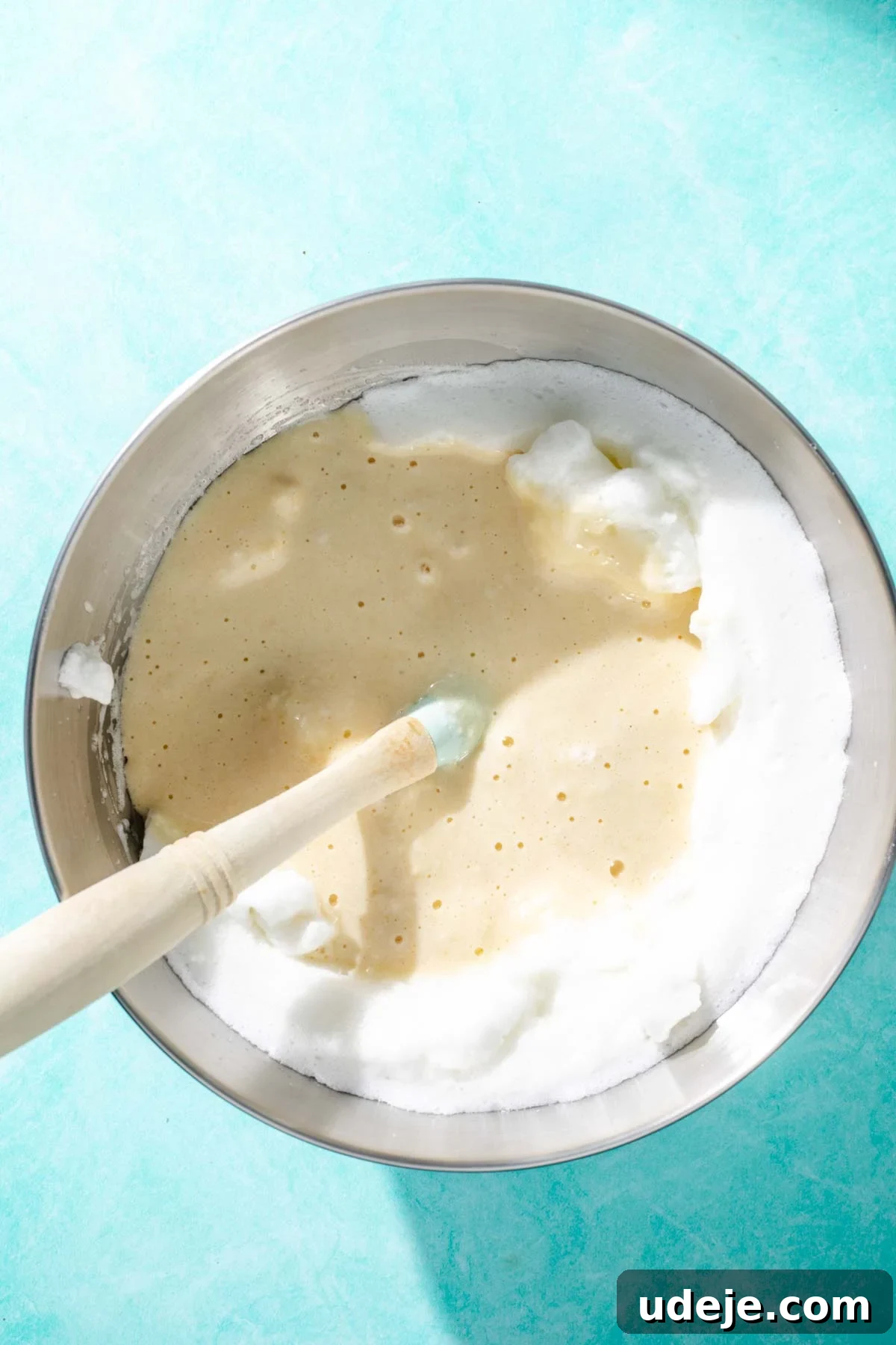 Heavenly Turkish Milk Cake 11 Egg whites being folded into cake batter with a blue spatula in a metal mixing bowl on a blue background