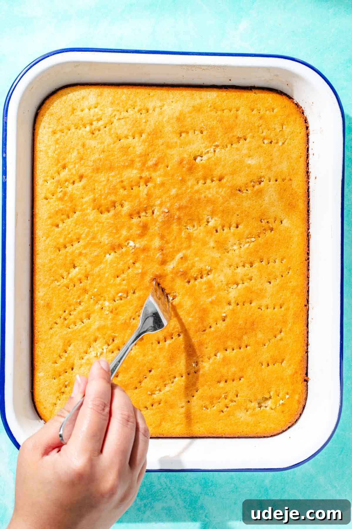 Heavenly Turkish Milk Cake 18 Holes being poked into cake by a fork in a white baking pan on a blue background