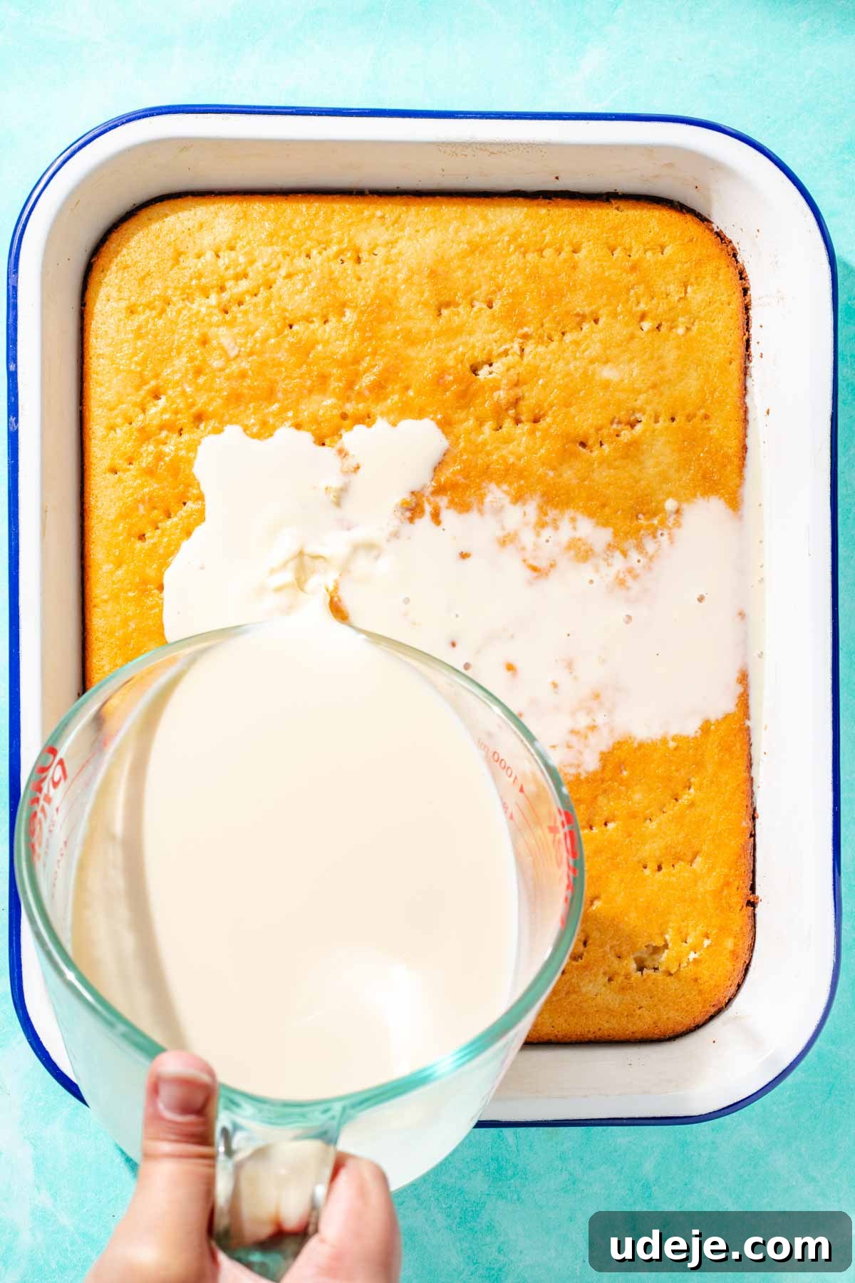 Heavenly Turkish Milk Cake 19 Milk mixture being poured from a glass measuring cup over cake in a white baking pan on a blue background