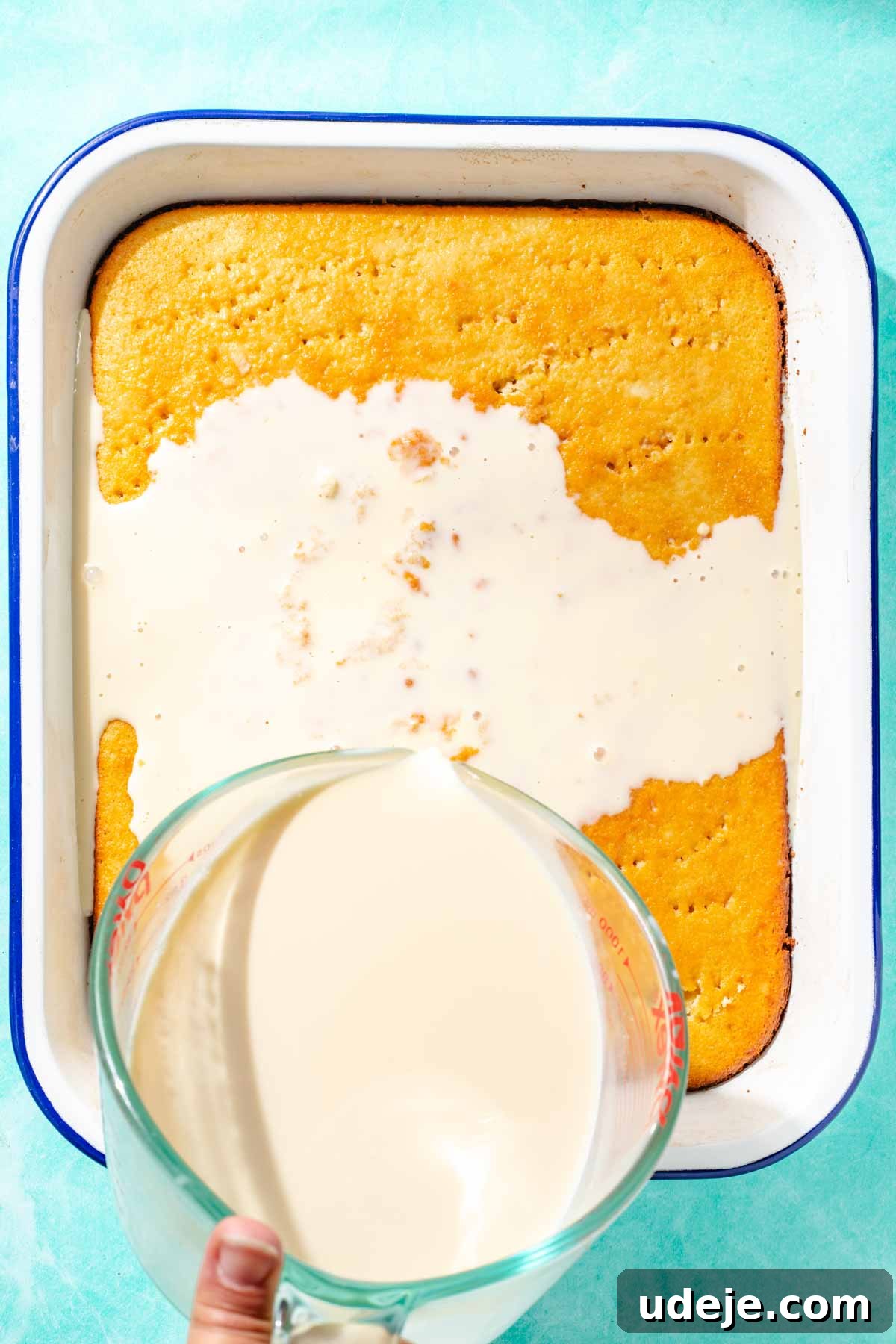 Heavenly Turkish Milk Cake 20 Milk mixture being poured from a glass measuring cup over cake in a white baking pan on a blue background