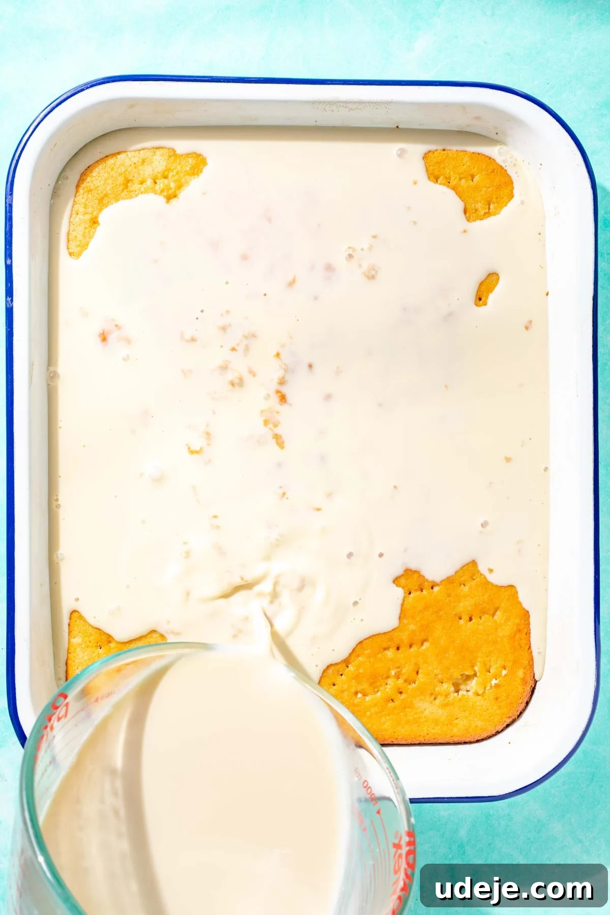 Heavenly Turkish Milk Cake 21 Milk mixture being poured from a glass measuring cup over cake in a white baking pan on a blue background