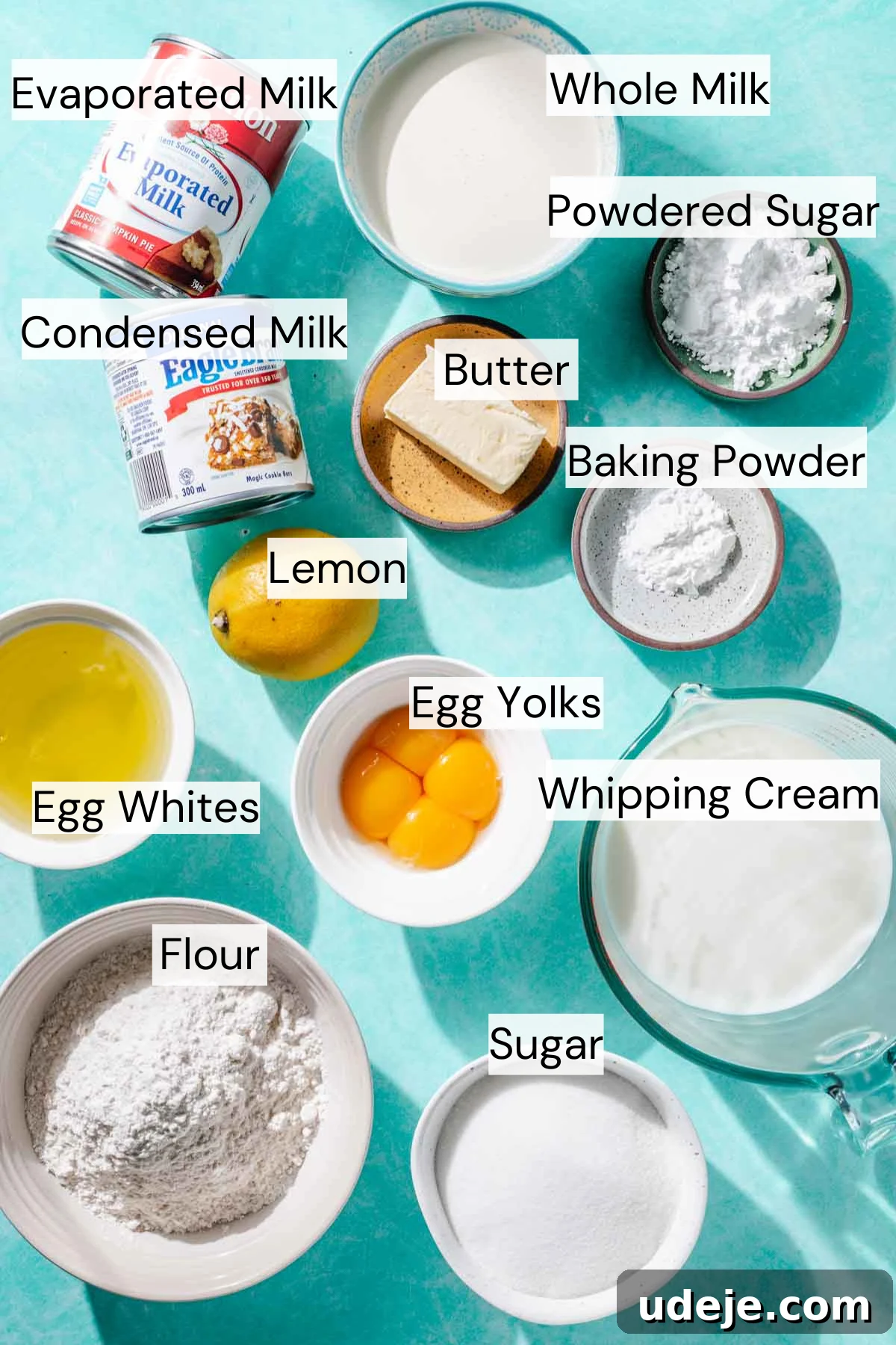 Heavenly Turkish Milk Cake 4 Ingredient shot for turkish tres leches with a bowl of whole milk, small bowl of powdered sugar, small bowl of baking powder, small bowl of butter, can of evaporated milk, can of condensed milk, lemon, egg whites in a bowl, egg yolks in a bowl, whipping cream in a glass measuring cup, bowl of flour and bowl of sugar on a blue background