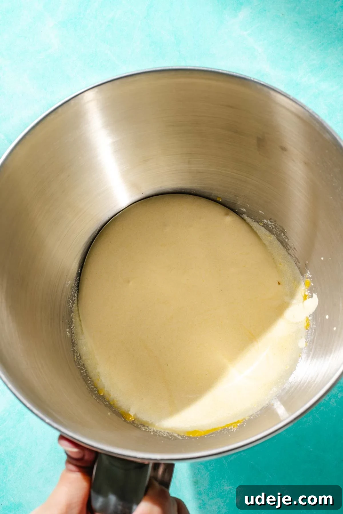 Heavenly Turkish Milk Cake 7 Metal mixing bowl with a hand holding the handle with light coloured whisked egg yolks on a blue background