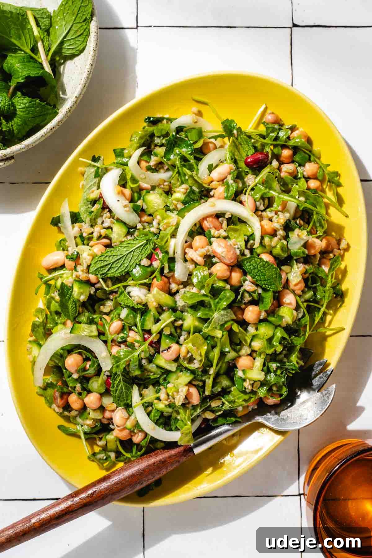 Silky Freekeh Bean Fiesta 2 A yellow oval platter with Freekeh Salad served inside. The salad includes arugula, chopped cucumber, sliced onions, beans, cooked freekeh, and chopped parsley. There is a small bowl with mint in the top left, and an orange water glass in the bottom right.