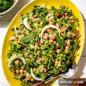 Silky Freekeh Bean Fiesta 12 A yellow oval platter with Freekeh Salad served inside. The salad includes arugula, chopped cucumber, sliced onions, beans, cooked freekeh, and chopped parsley. There is a small bowl with mint in the top left, and an orange water glass in the bottom right.