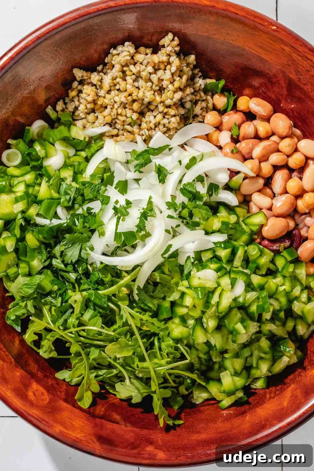 Silky Freekeh Bean Fiesta 8 A large wooden salad bowl with the following ingredients inside: arugula, chopped cucumber, sliced onions, beans, cooked freekeh, and chopped parsley