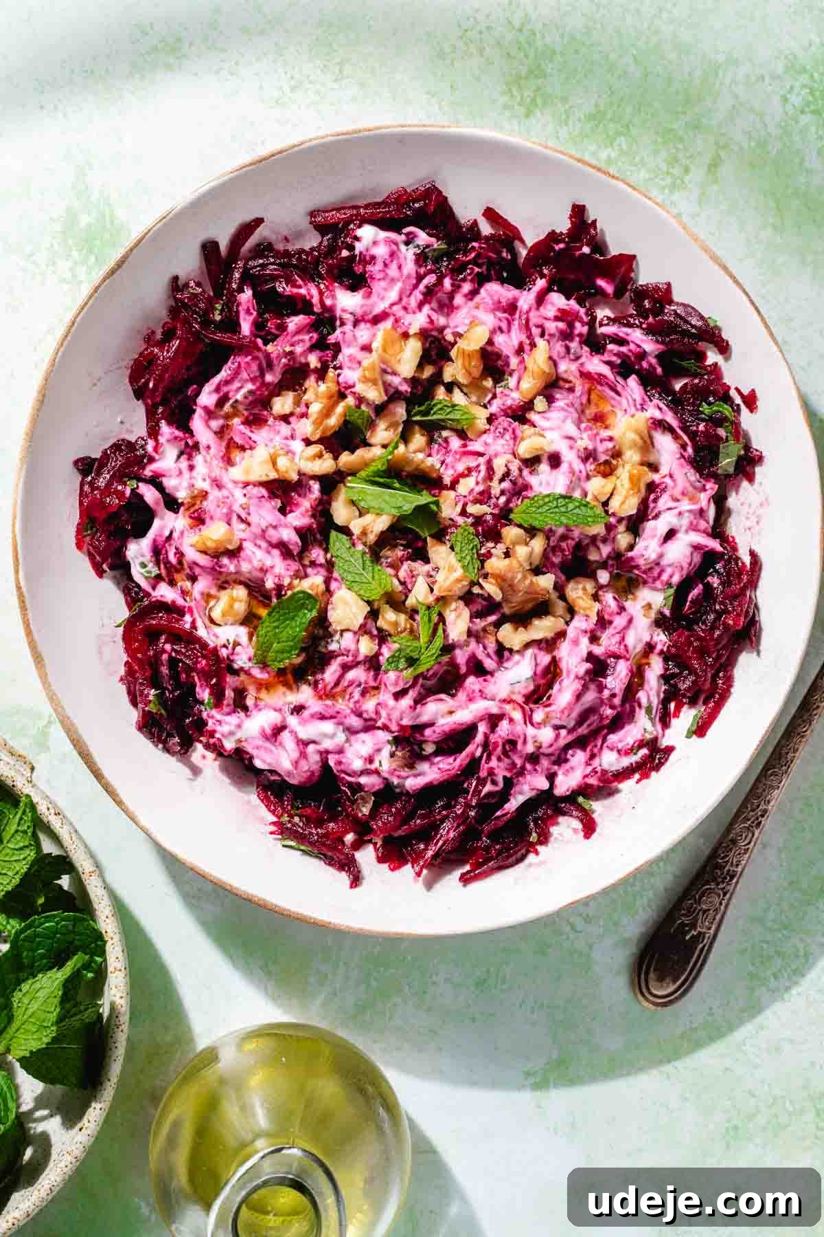 Vibrant Beetroot Yogurt Salad 2 Turkish beet salad garnished with walnuts and mint in a white salad bowl on a green background with a fork at the side, a small beige bowl with mint leaves and a small oil dispenser with olive oil