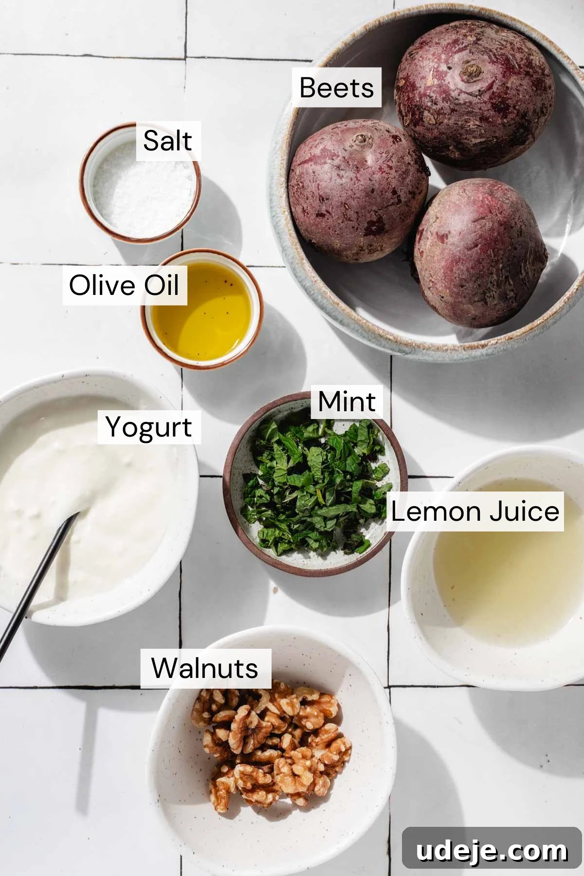 Vibrant Beetroot Yogurt Salad 3 Ingredient shot for Turkish Beet Salad with a bowl with 3 whole beets, a small bowl of salt, a small bowl of olive oil, a bowl of mint leaves, a bowl of yogurt with a spoon, a bowl of lemon juice and a bowl of walnuts on a white tiled background