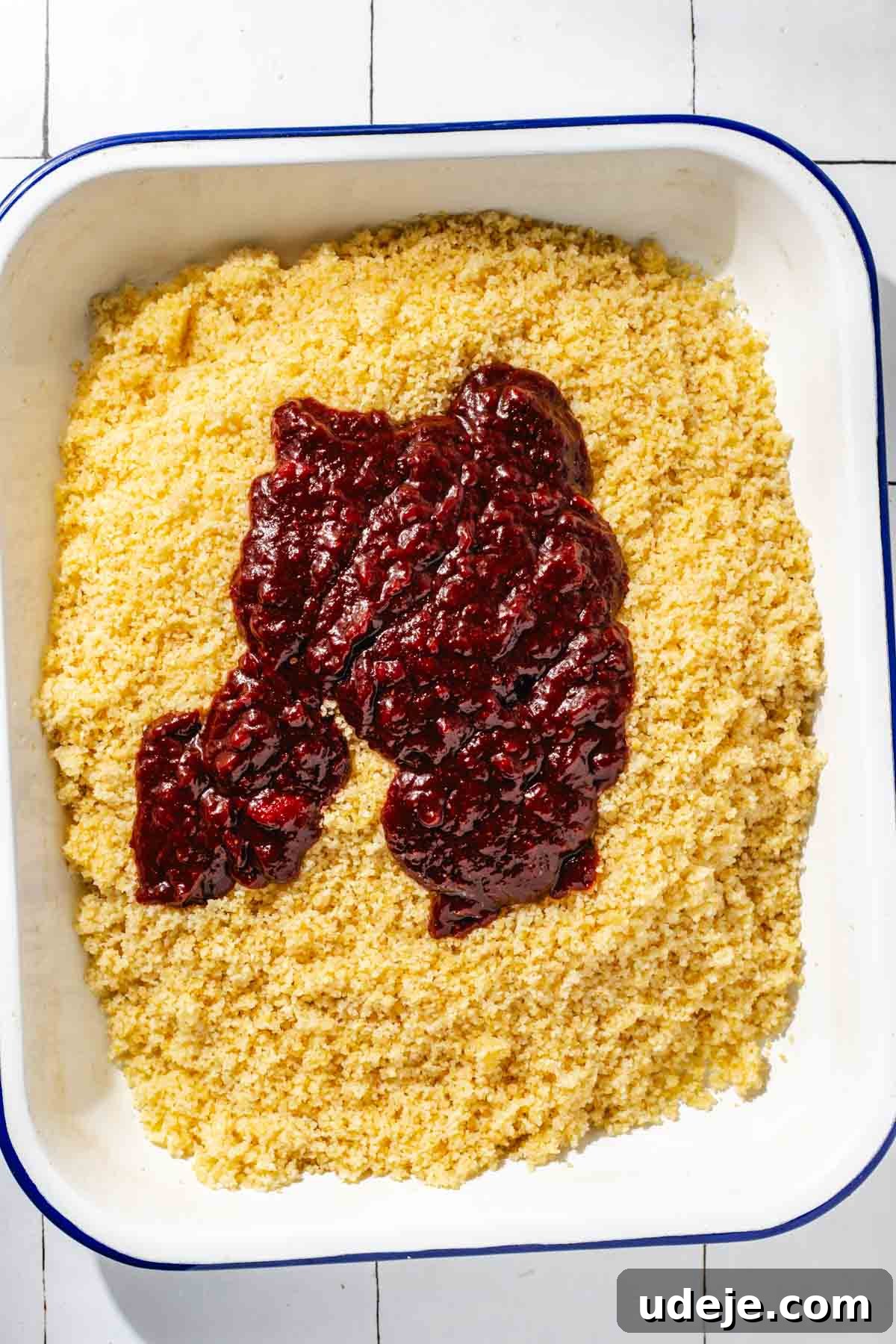 Fast 20-Minute Turkish Bulgur Kisir 11 The vibrant red Kisir dressing poured over the fluffed bulgur in a white, blue-rimmed baking pan, signaling the next step in preparing this Turkish salad, on a white tiled surface.