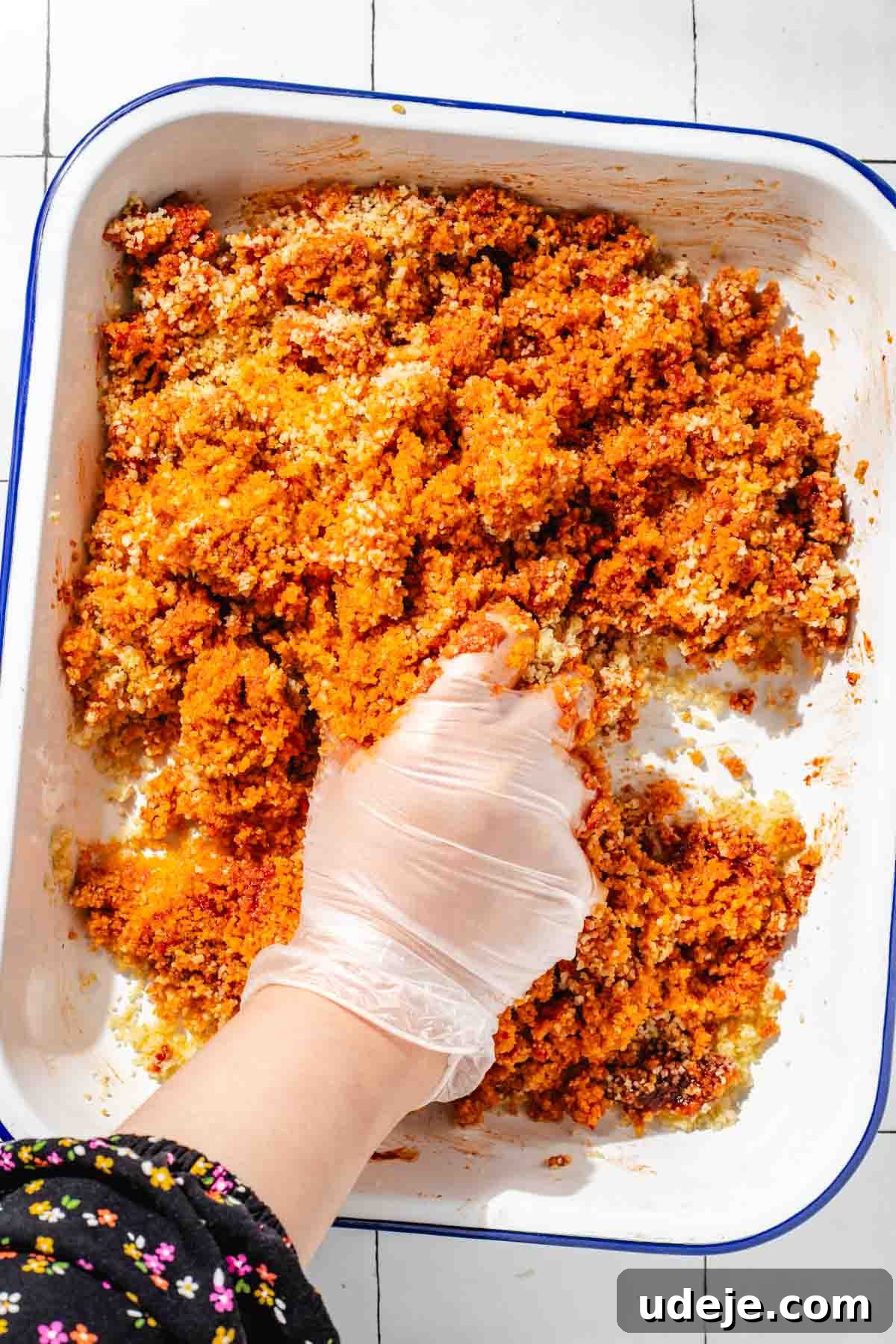 Fast 20-Minute Turkish Bulgur Kisir 12 Hands expertly mixing the bulgur and rich red Kisir dressing in a white, blue-rimmed baking pan, demonstrating the traditional rubbing technique on a white tiled surface.