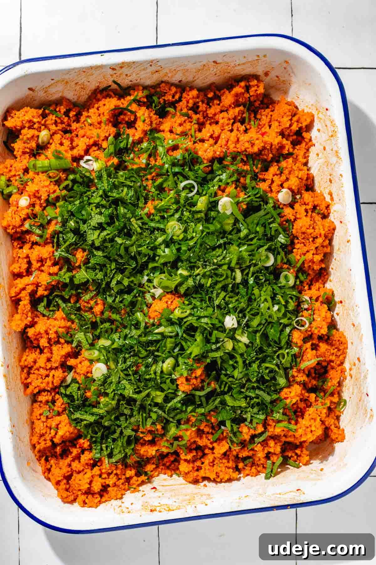 Fast 20-Minute Turkish Bulgur Kisir 13 The vibrant Kisir bulgur mix with a generous scattering of fresh green herbs and chopped green onions on top, awaiting final integration in a white, blue-rimmed baking pan on a white tiled surface.