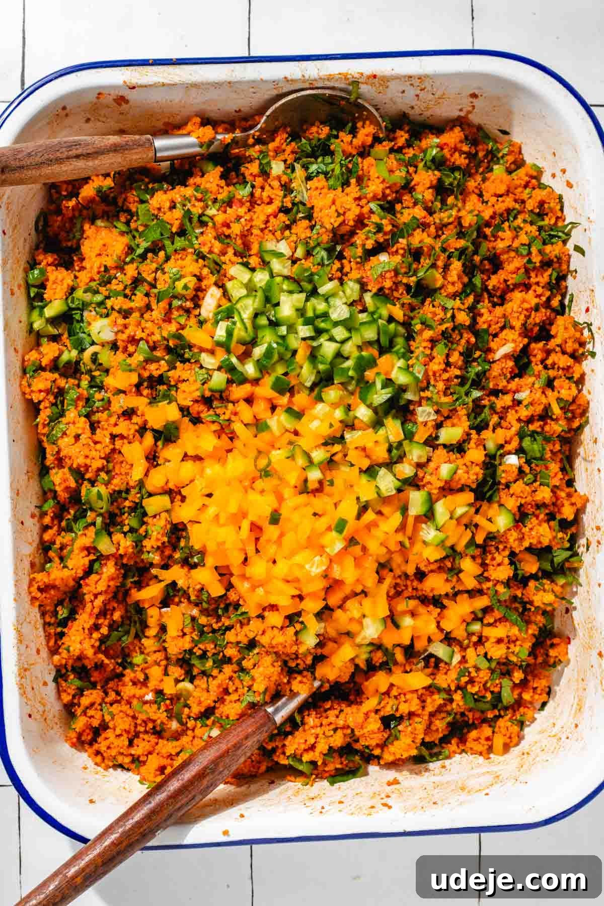 Fast 20-Minute Turkish Bulgur Kisir 14 Fully mixed Kisir, vibrant red bulgur dotted with finely chopped cucumbers and bell peppers, presented in a white, blue-rimmed baking tray with two elegant wooden serving utensils on the sides, on a white tiled surface.